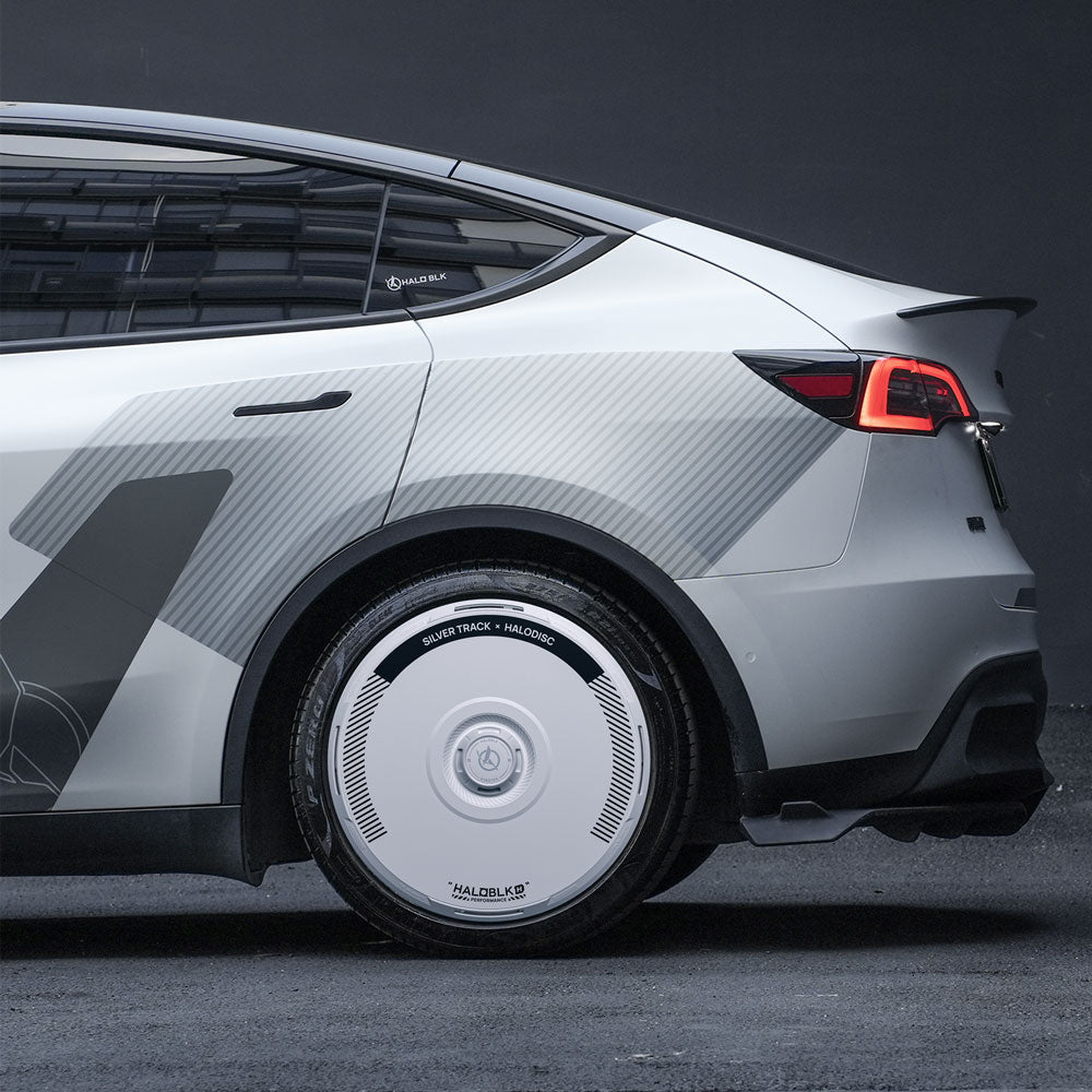A close-up side view of a Tesla Model Y with HALOBLK Store’s HALODISC 2 Wheel Covers in Silver Track, designed for 19" Gemini wheels, featuring unique aerodynamic covers and geometric decals, parked on a dark surface against a dark background.