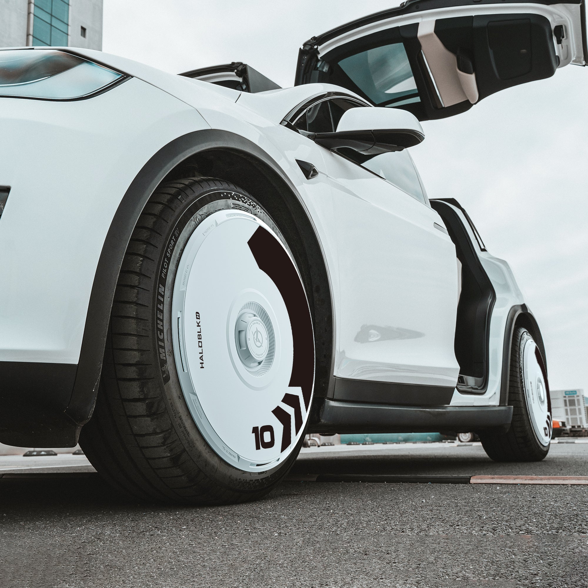 A white Tesla Model X (2021-2025) with its gullwing door open features a customized HALODISC 2 Wheel Cover by HALOBLK Store on its 20" Perihelix Wheel, showcasing a unique white hubcap with black detailing and the number 10.