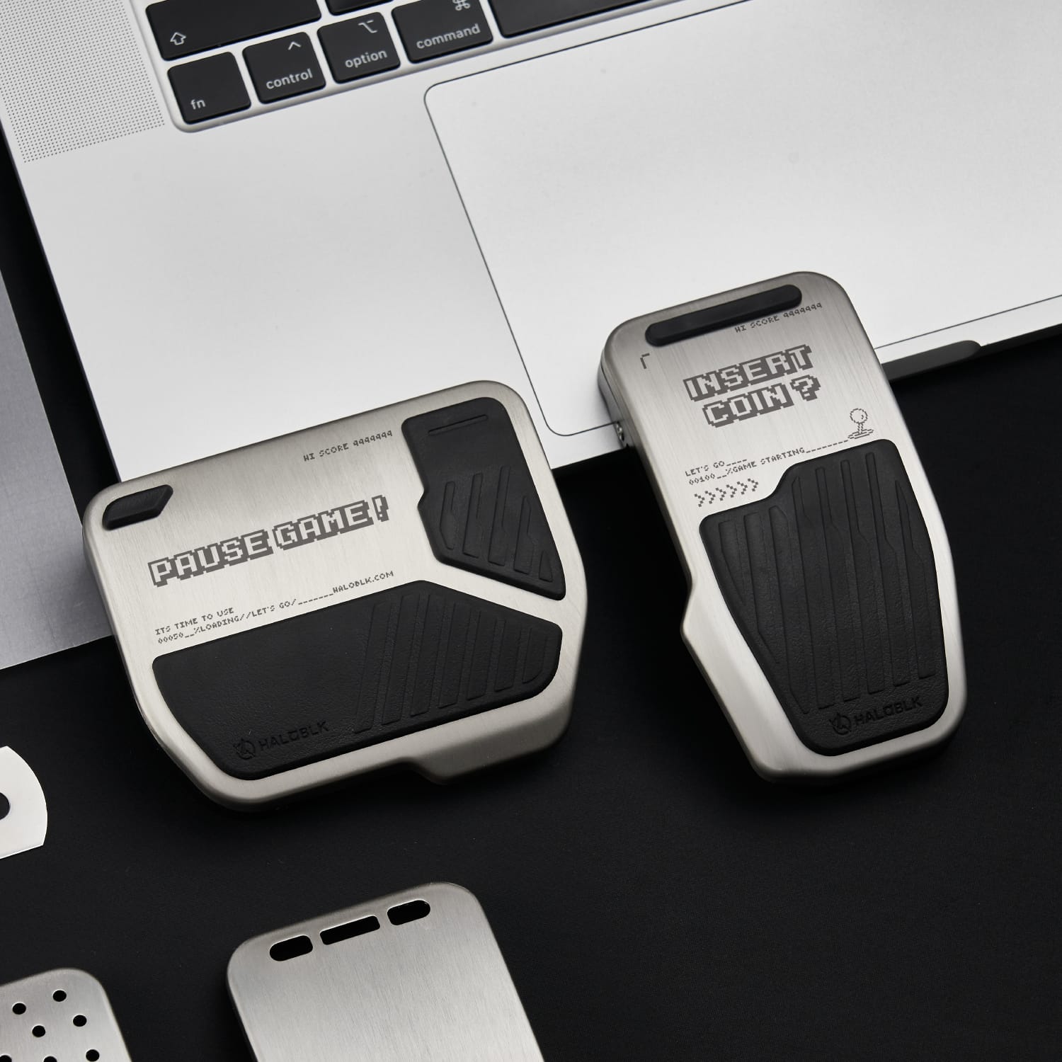 Two Alloy Pedals 2 for Tesla by HALOBLK Store, each with black rubber grips and arcade-themed text, feature a sleek, industrial design and sit beside a laptop on a black surface.