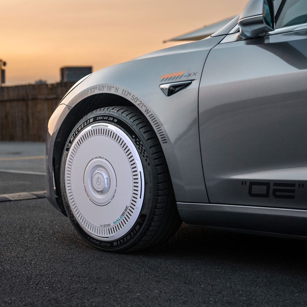 Close-up of a Tesla Model 3 2017-2018 fitted with HALODISC 2 Wheel Covers from HALOBLK Store, featuring a unique white and silver design on 19" performance wheels. Bold white tire lettering is visible as the car is parked at sunset.