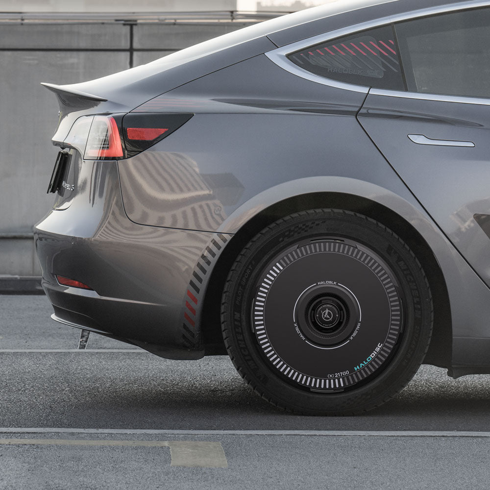 A close-up side view of a Tesla Model 3 (2017-2018) with HALODISC 2 Wheel Covers from HALOBLK Store on 19" Performance wheels, showing their futuristic circular patterns; a concrete barrier and urban street appear in the background.