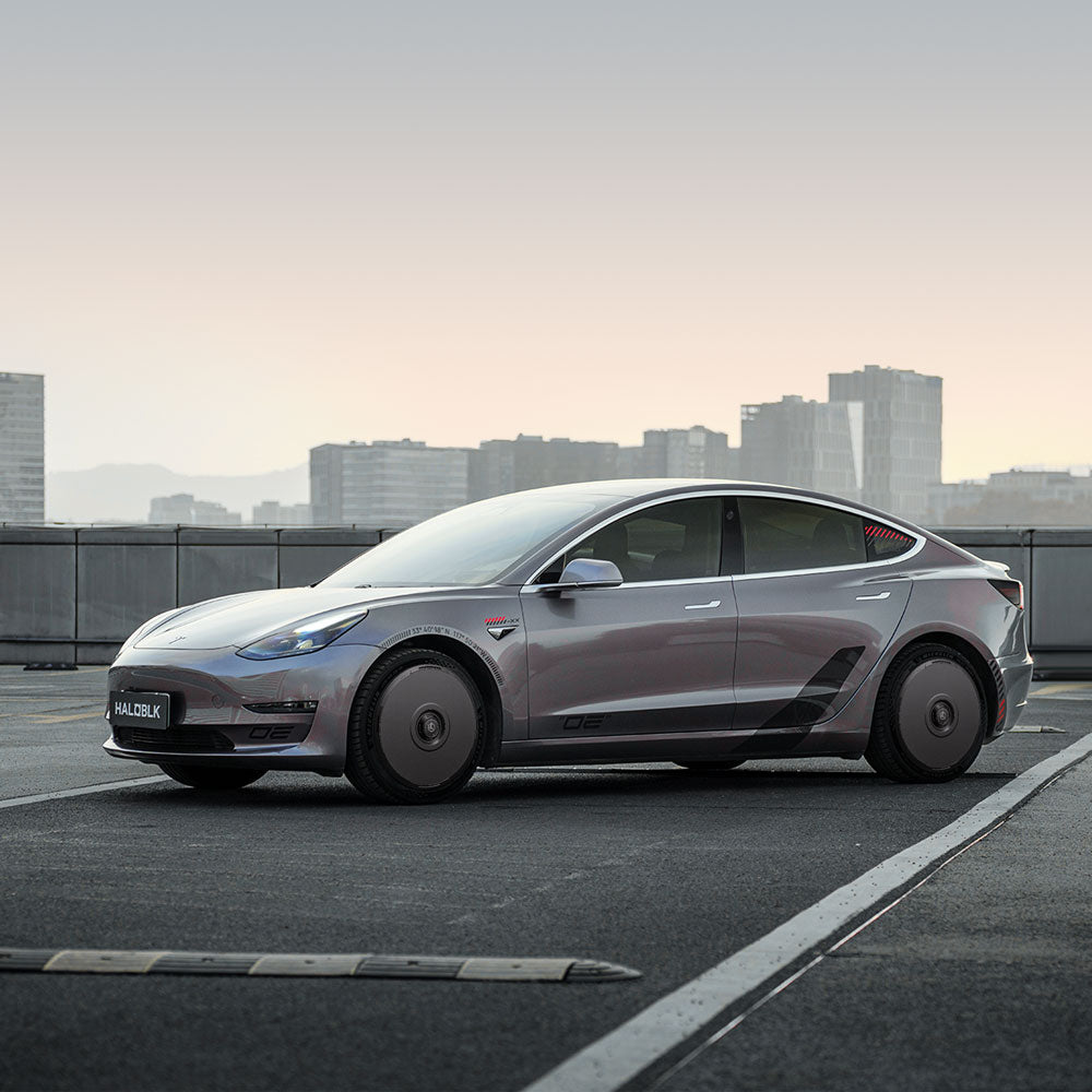 A silver Tesla Model 3 (2019-2023) with HALODISC 2 Wheel Covers by HALOBLK Store is parked on a rooftop at dusk, sleek lines and dark 19" sport wheels set against a city skyline.