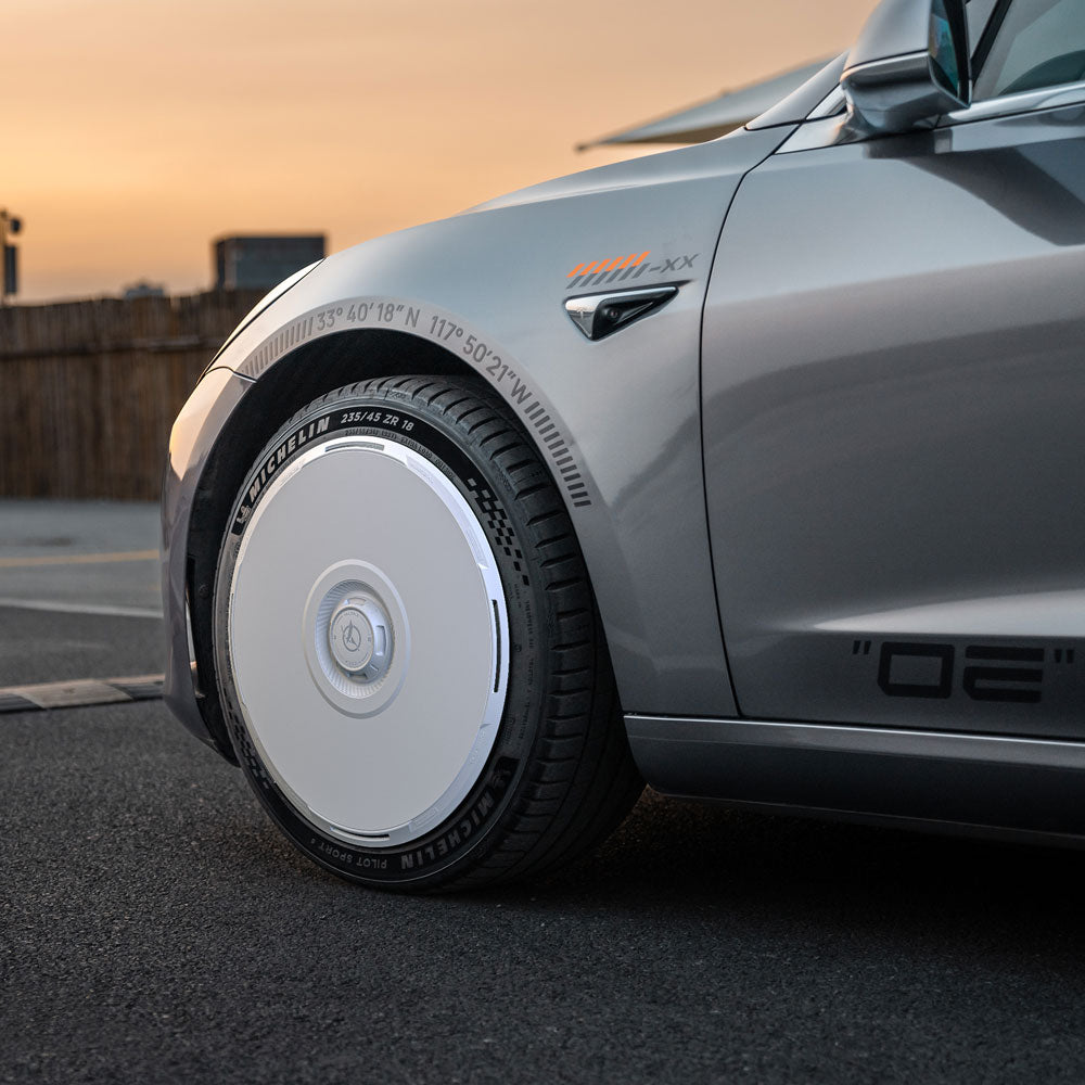 Close-up of a silver car parked at sunset, showcasing HALODISC 2 Wheel Covers for Tesla Model 3 2018-2023 19" Stiletto Wheels by HALOBLK Store. Features white aerodynamic covers and visible front side details.