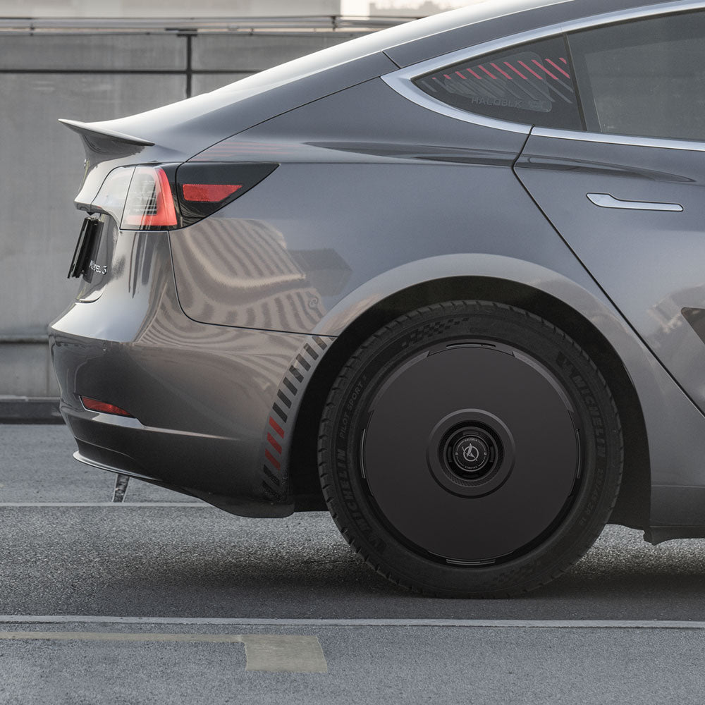 A close-up of a gray Tesla Model 3 (2018-2023) with sleek design, parked by a concrete barrier, showcases the unusual solid black HALODISC 2 Wheel Covers for 19" Stiletto Wheels from HALOBLK Store.