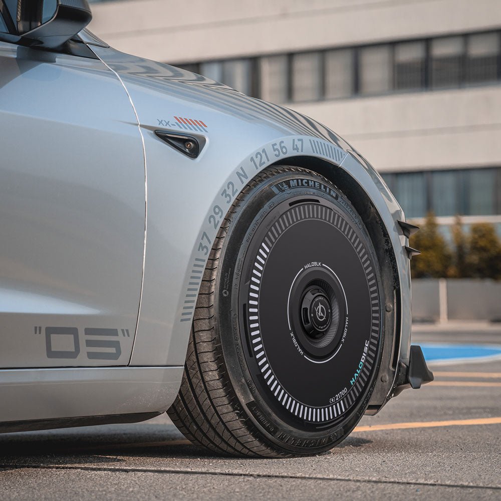 Close-up of a Tesla Model 3 Highland with HALODISC 2 Wheel Covers by HALOBLK Store on its 18" Photon Wheels, showing their unique futuristic black design with numbers and markings, parked in front of a modern building.