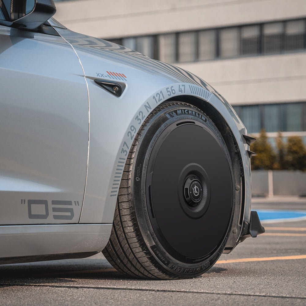 A close-up of a Tesla Model 3 Highland with HALODISC 2 Wheel Covers (18" Photon) from HALOBLK Store, featuring a futuristic black design with coordinates and numbers, parked near a modern building.