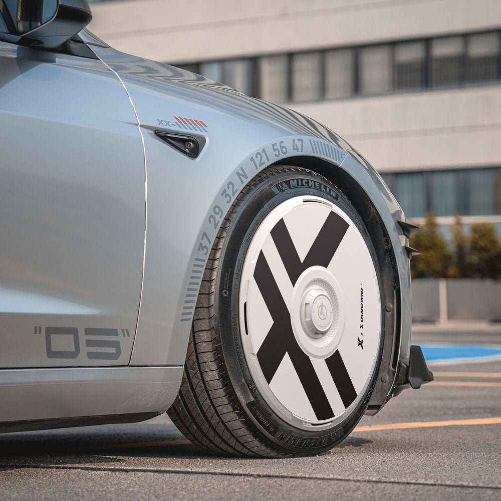A close-up of HALODISC 2 Wheel Covers for Tesla Model 3 Highland 19" Nova Wheels by HALOBLK Store, showing the silver car’s front wheel with white X-Factor hubcaps and numbered markings, parked outside a modern building.
