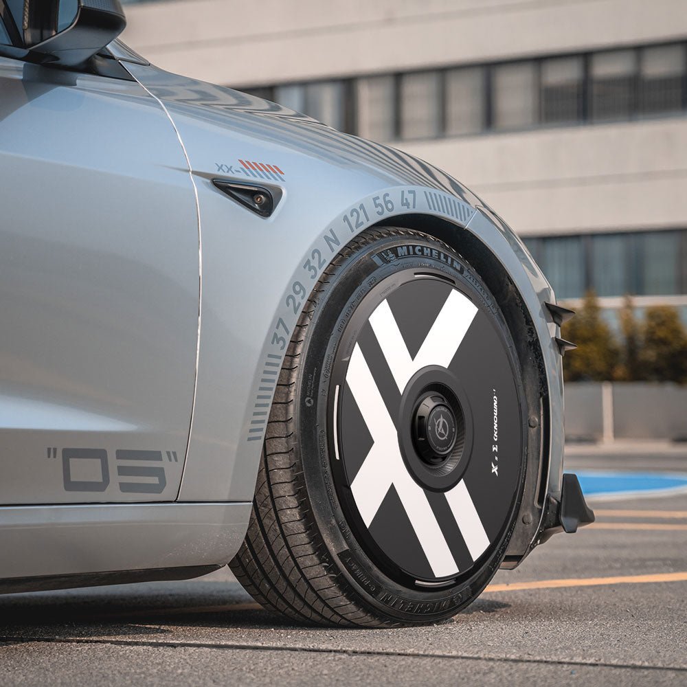 Close-up of a Tesla Model 3 Highland with HALODISC 2 Wheel Covers (X-Factor) by HALOBLK, featuring bold white X-shaped accents and numbered details on black covers, parked by a modern building. Suitable for 19" Nova wheels.