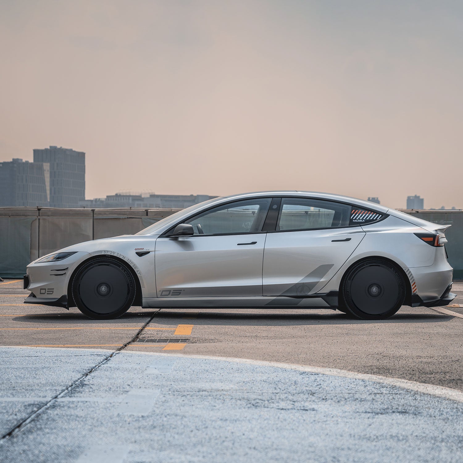 A Tesla Model 3 Highland with HALODISC 2 Wheel Covers for 20" Warp Wheels from HALOBLK Store is parked on a rooftop, city buildings in the background beneath an overcast sky.