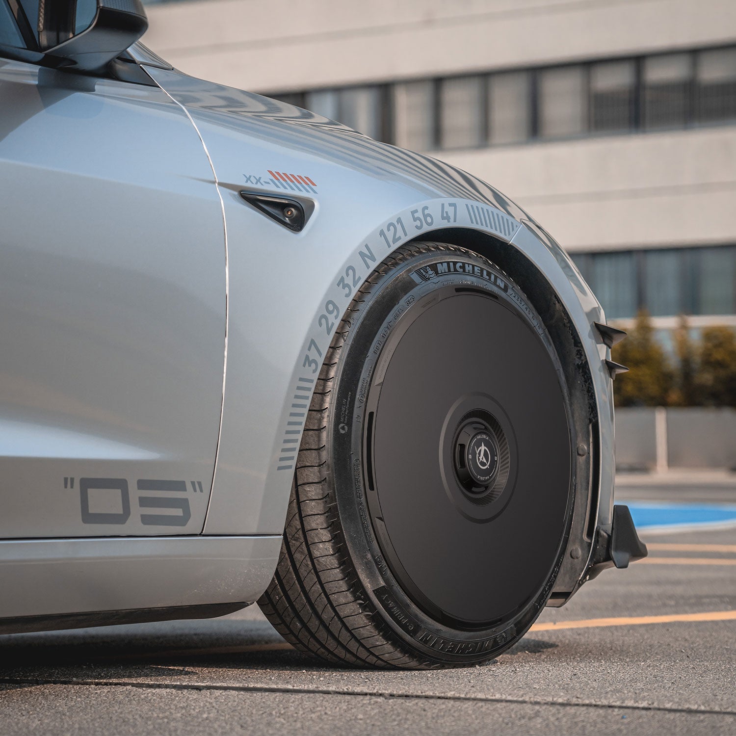 Close-up of HALODISC 2 Wheel Covers for Tesla Model 3 Highland 20" Warp Wheels by HALOBLK Store, in sleek silver with black, aerodynamic design and numbered wheel arch. Modern building and hedge appear in the background.