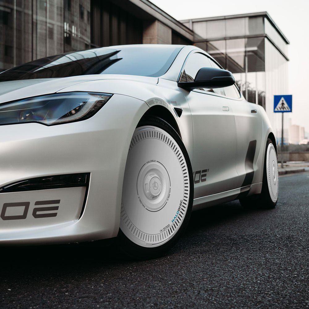 A Tesla Model S with HALODISC 2 Wheel Covers by HALOBLK Store is parked on a city street near a glass building and a pedestrian crossing sign.