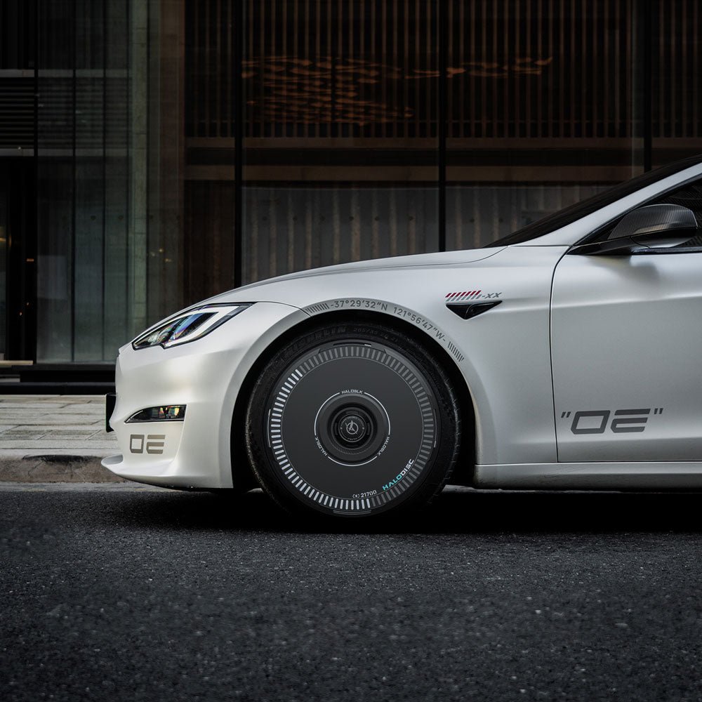 A white Tesla Model S with HALODISC 2 Wheel Covers by HALOBLK Store is parked in front of a modern glass building, displaying custom graphics and futuristic styling. Only the front half of the car is visible.