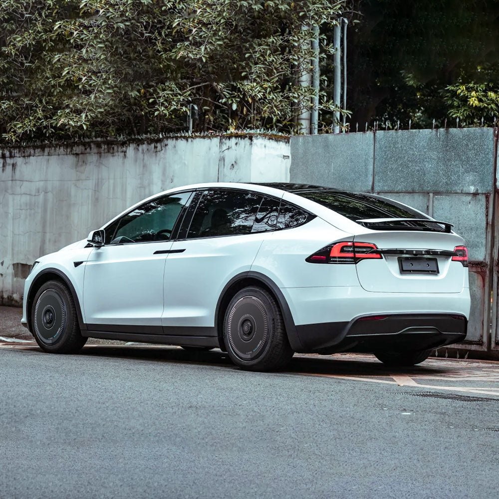 A white Tesla Model X (2016-2020) with HALODISC 2 Wheel Covers by HALOBLK Store on its 20" Slipstream wheels is parked by a street next to a concrete wall, with greenery and trees in the background; rear and side views are visible.