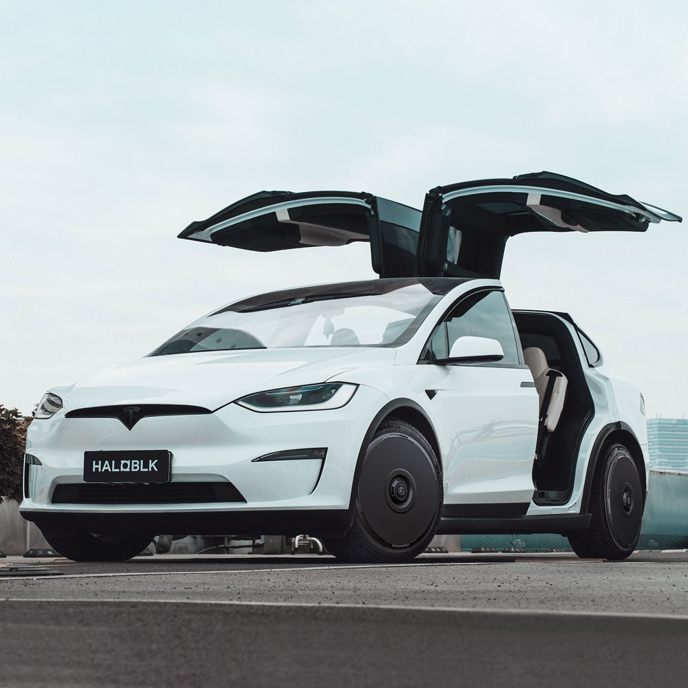 A white Tesla Model X (2016-2020) with HALODISC 2 Wheel Covers by HALOBLK Store, falcon-wing doors open, parked on a city street under a cloudy sky. The license plate reads HALOBLK.