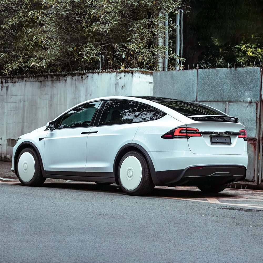 A white Tesla Model X featuring HALODISC 2 Wheel Covers for 2016-2020 20" Slipstream Wheels from the HALOBLK Store is parked by a tall gray wall with greenery in the background.