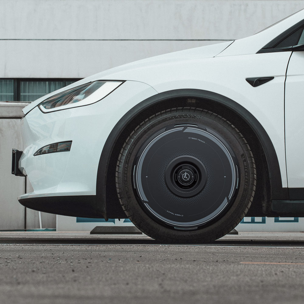 A close-up side view of a white Tesla Model X (2021-2025) shows its front wheel fitted with HALODISC 2 Wheel Covers from HALOBLK Store, featuring a sleek black and silver design. Part of a building and pavement are visible in the background.