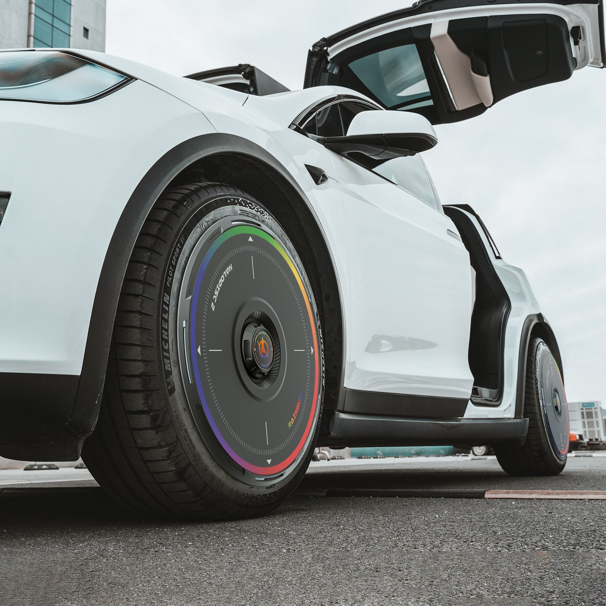 A white Tesla Model X (2016-2020) with a gull-wing door open is parked, showcasing the HALODISC 2 "Rainbow Road" wheel covers by HALOBLK Store on its front 20" Slipstream wheel. Buildings and a cloudy sky appear in the background.