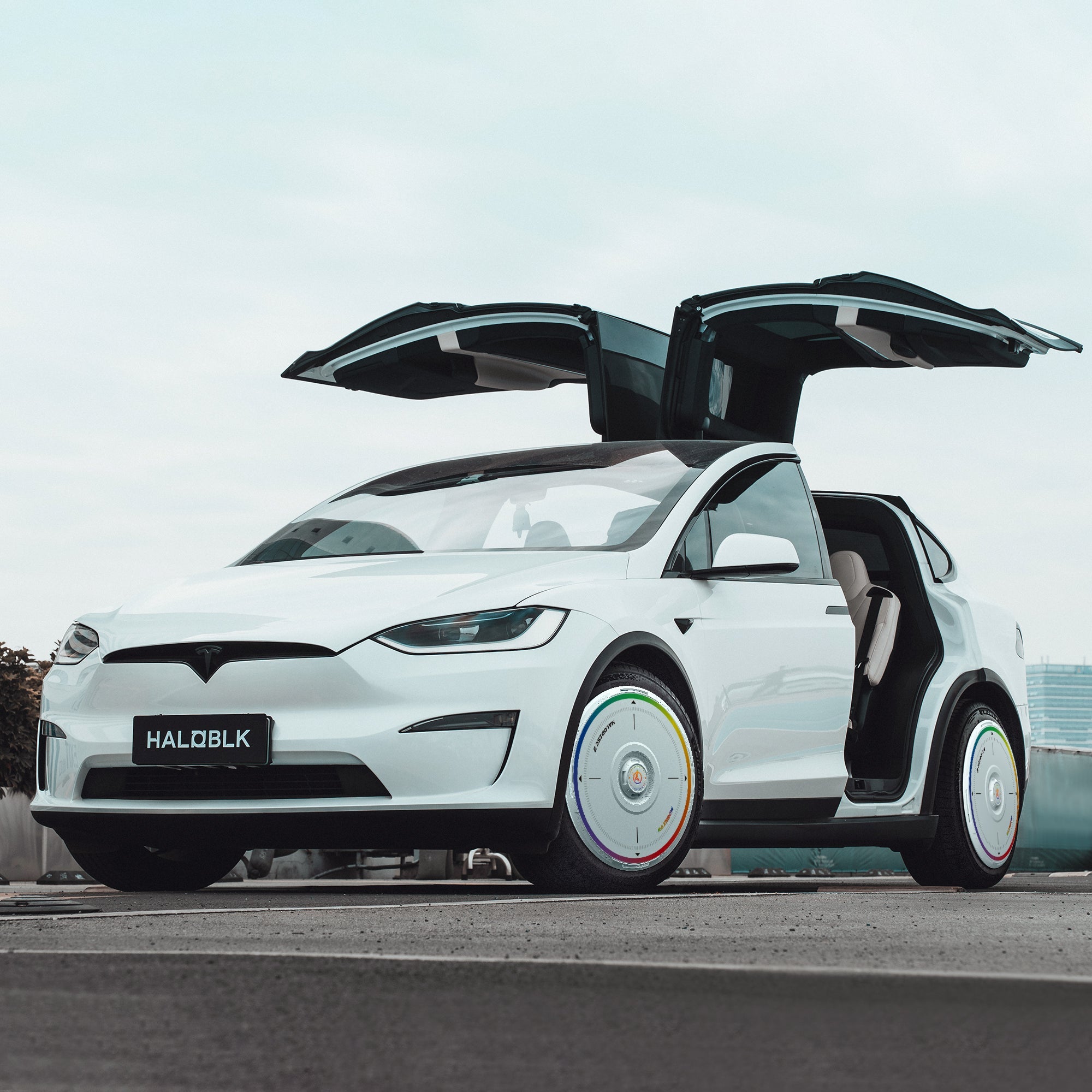 A white Tesla Model X (2016-2020) with HALODISC 2 Rainbow Road wheel covers from HALOBLK Store is parked on a rooftop, falcon-wing doors open, highlighting its futuristic design.