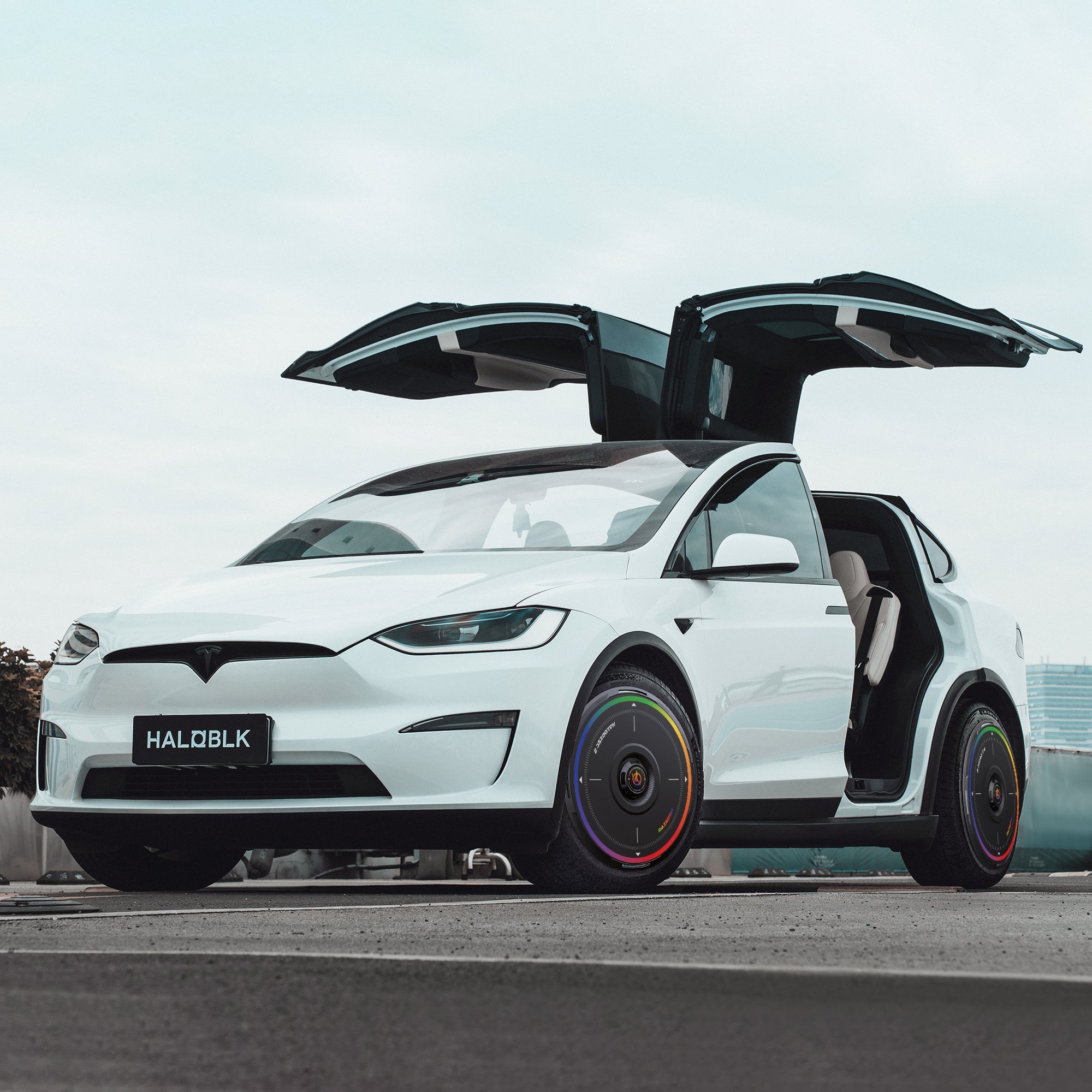 A white Tesla Model X (2016-2020) with gull-wing doors open, parked on a city street, features colorful HALODISC 2 Rainbow Road wheel covers by HALOBLK Store and a license plate reading HALOBLK.
