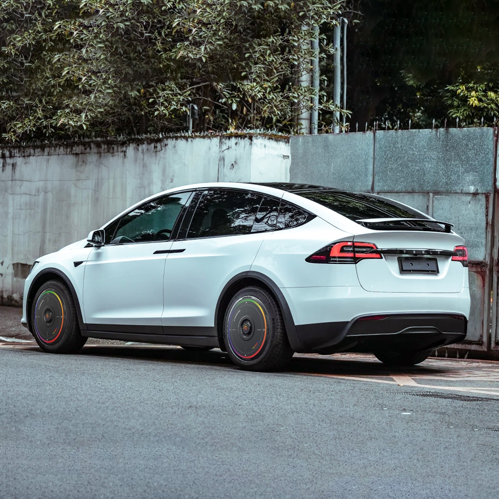 A white Tesla Model X (2016-2020) with HALODISC 2 Rainbow Road wheel covers from HALOBLK Store is parked by a street, with trees and a tall concrete wall in the background.