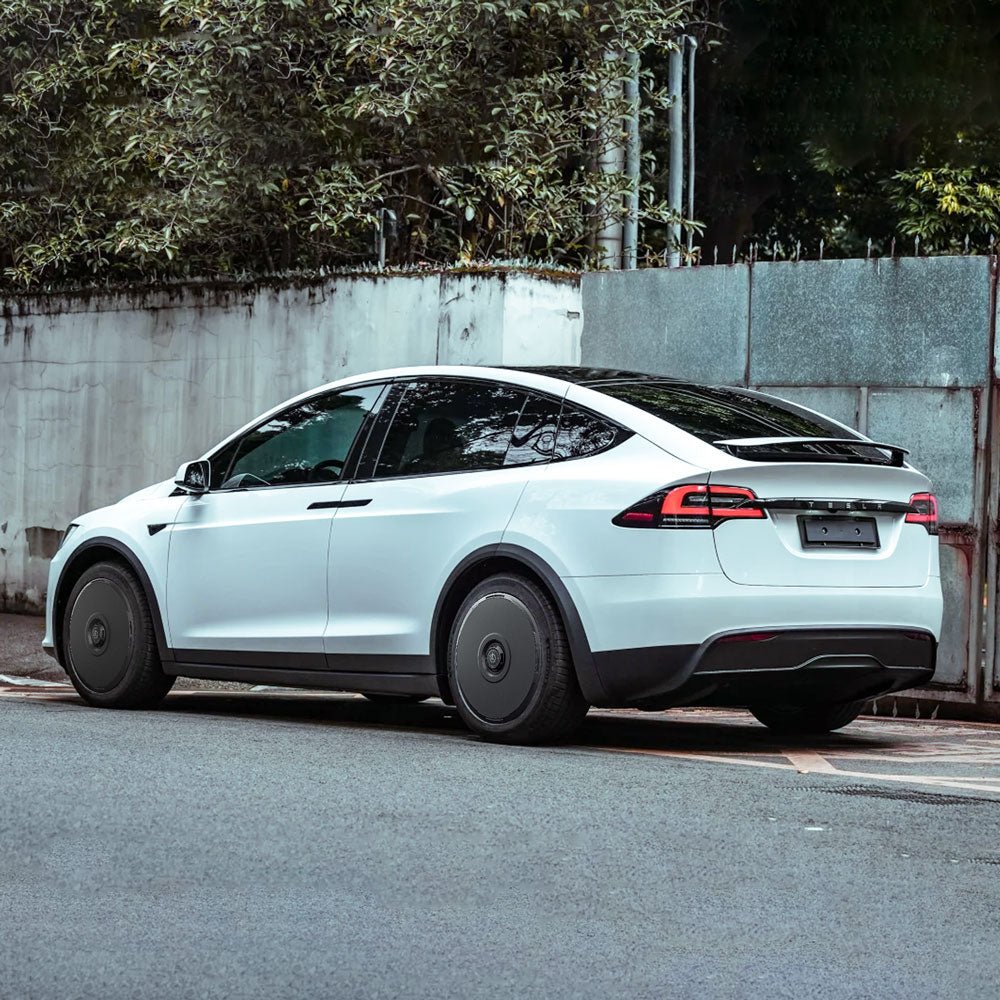 A white Tesla Model X (2021-2025) with HALODISC 2 Wheel Covers by HALOBLK Store is parked on a street near a concrete wall with greenery and fencing, featuring tinted windows and stylish dark 20" Cyberstream wheels.