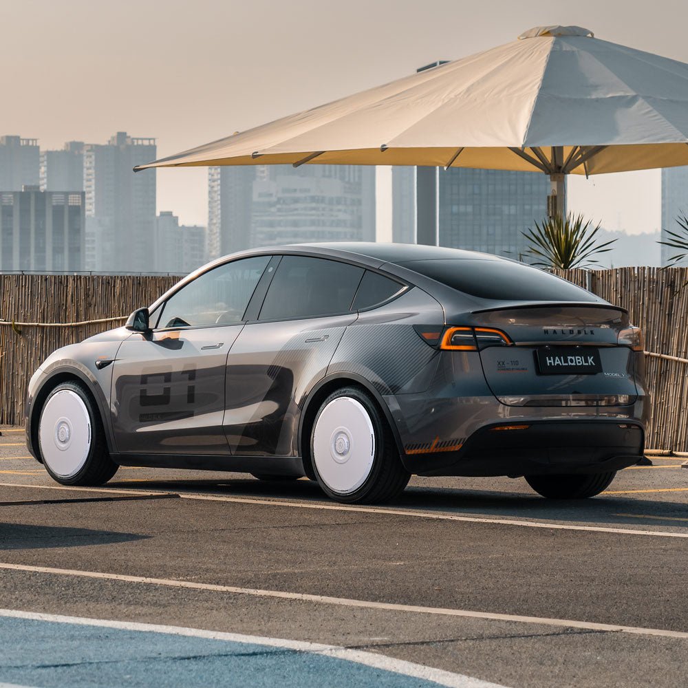 A Tesla Model Y with HALODISC 2 Wheel Covers by HALOBLK Store is parked outdoors near a large umbrella, with a city skyline and tall buildings in the background.