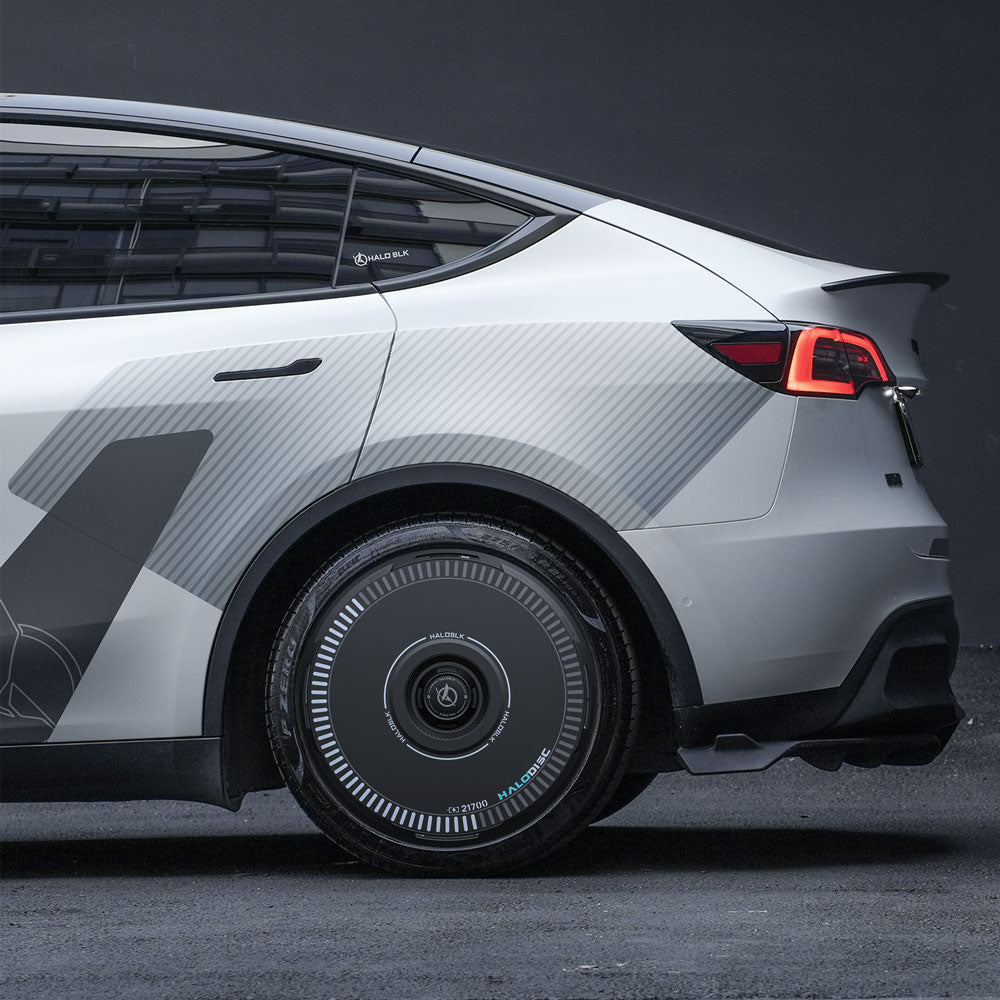 A close-up side view of a silver Tesla Model Y with HALODISC 2 Wheel Covers from HALOBLK Store on 21" Überturbine Wheels, showcasing geometric patterns and unique black wheels with blue accents, parked on a dark surface against a plain backdrop.