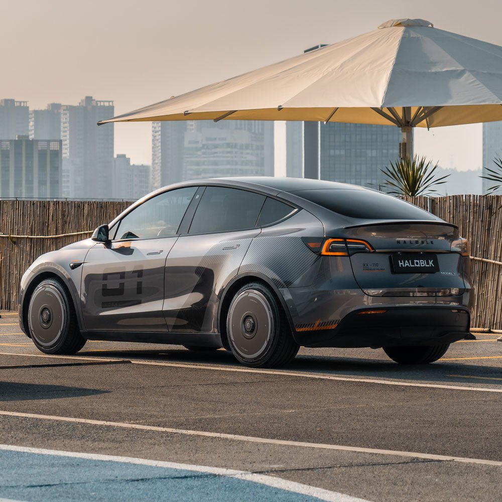 A Tesla Model Y with HALODISC 2 Wheel Covers by HALOBLK Store is parked near a bamboo fence under a large cream umbrella, with city buildings in the background. The sleek, futuristic covers fit 21" Überturbine wheels.
