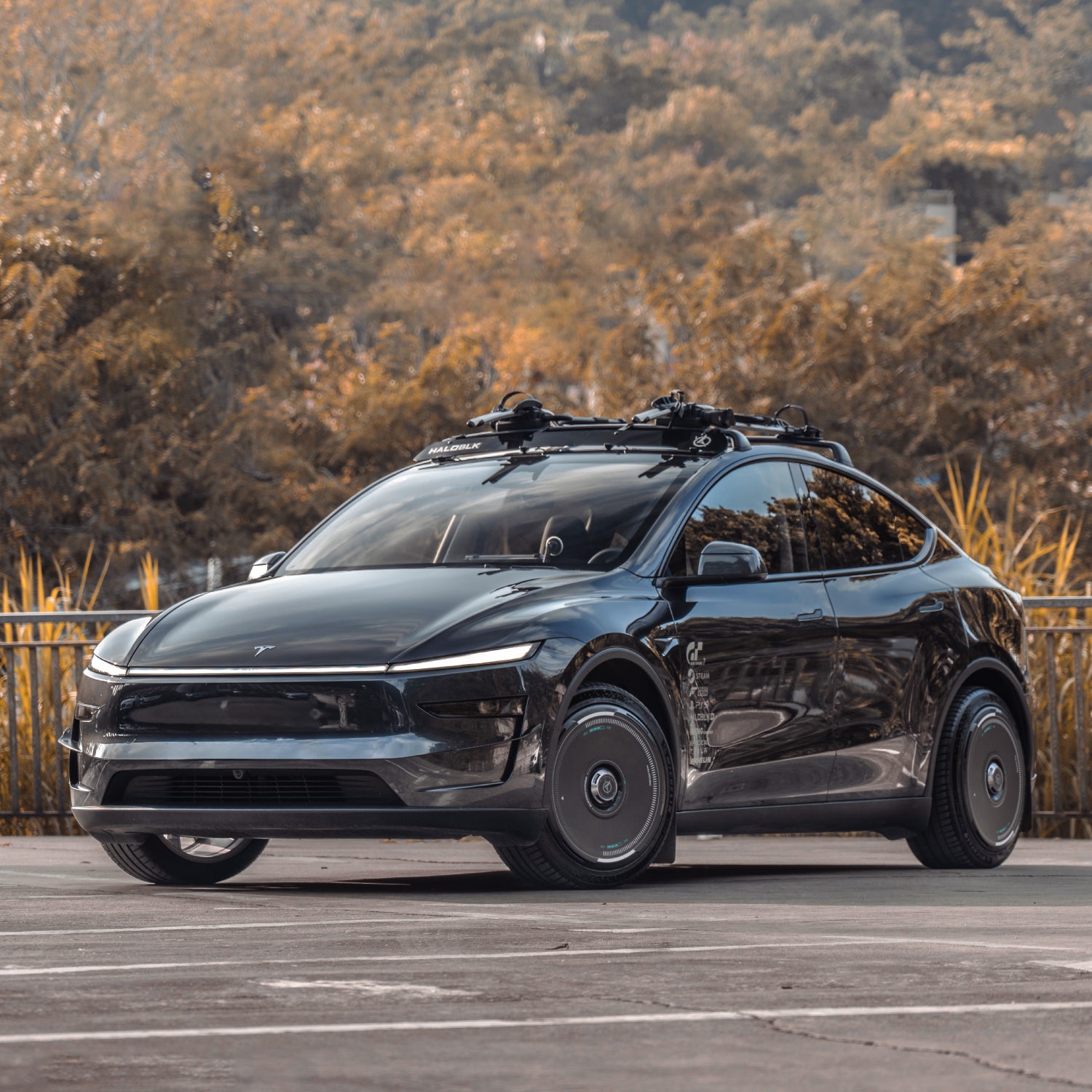 A sleek black Tesla Model Y featuring HALODISC 2 Wheel Covers by HALOBLK Store is parked outdoors in front of autumn trees, equipped with a roof rack and angled slightly to the right.