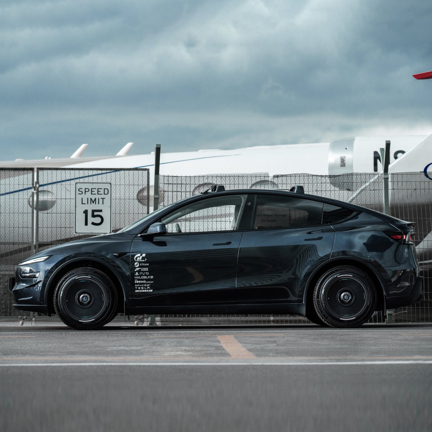 A sleek black Tesla Model Y with HALODISC 2 Wheel Covers by HALOBLK Store is parked on pavement near an airport fence, a speed limit 15 sign, and part of an airplane under a cloudy sky.