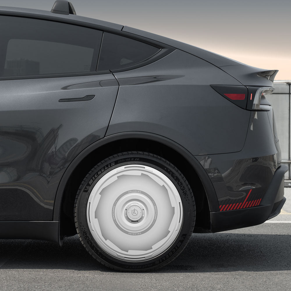 A close-up of a Tesla Model Y parked on a city street, featuring HALODISC 2 Wheel Covers by HALOBLK Store, highlighting the unique white, geometric Vision-GT design on the 19" Juniper Crossflow wheels.