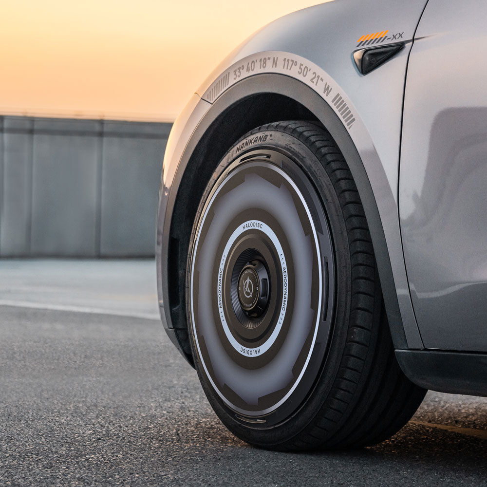 Close-up of HALODISC 2 Wheel Covers for Tesla Model Y 19" Gemini Wheels by HALOBLK Store, featuring a sleek Vision-GT hubcap design and unique fender markings, set outdoors at sunset.