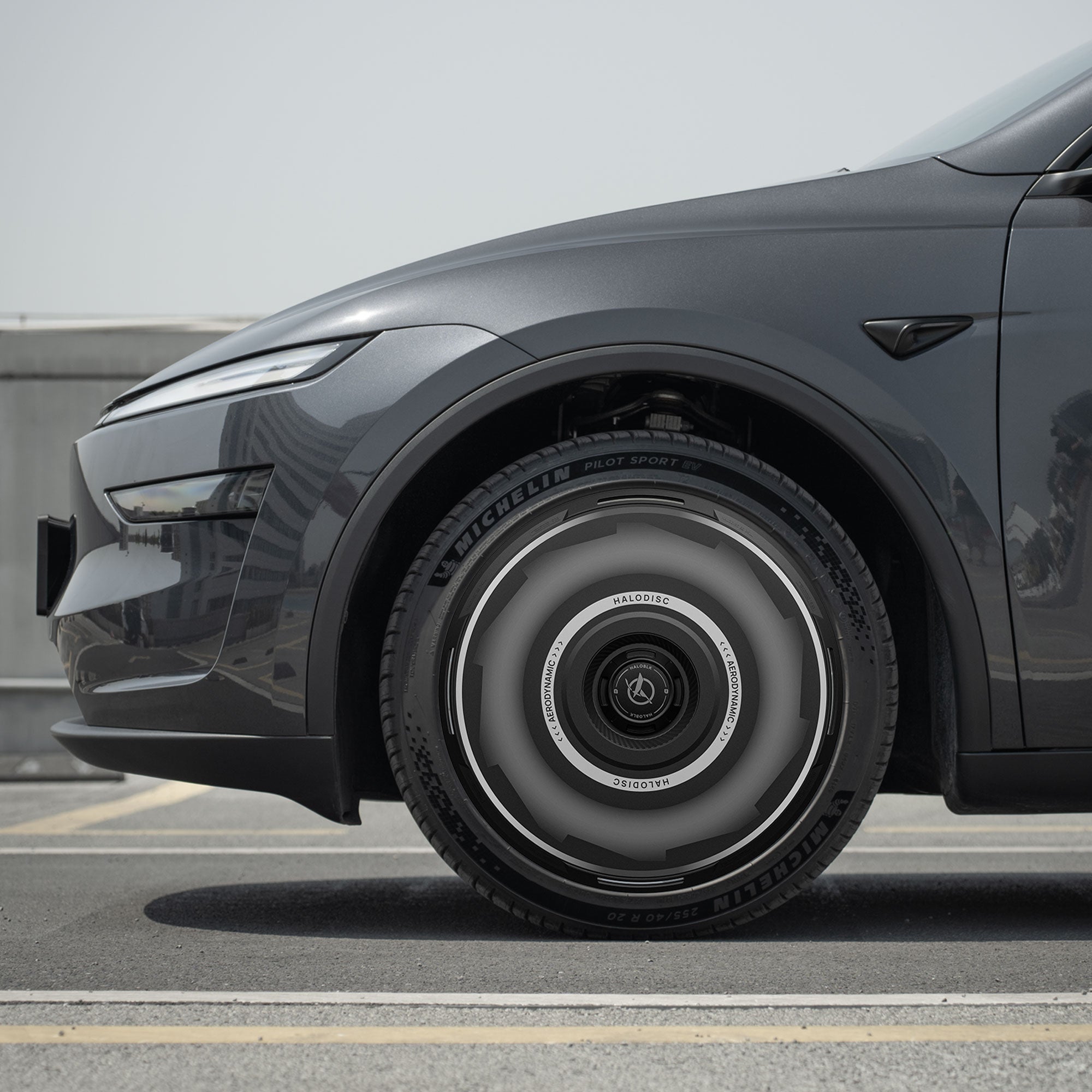 Showcasing the HALODISC 2 Wheel Covers for Tesla Model Y Juniper 19" Crossflow Wheels by HALOBLK Store—a sleek, dark gray car with futuristic, metallic Vision-GT wheel covers, parked on a rooftop under a clear sky.