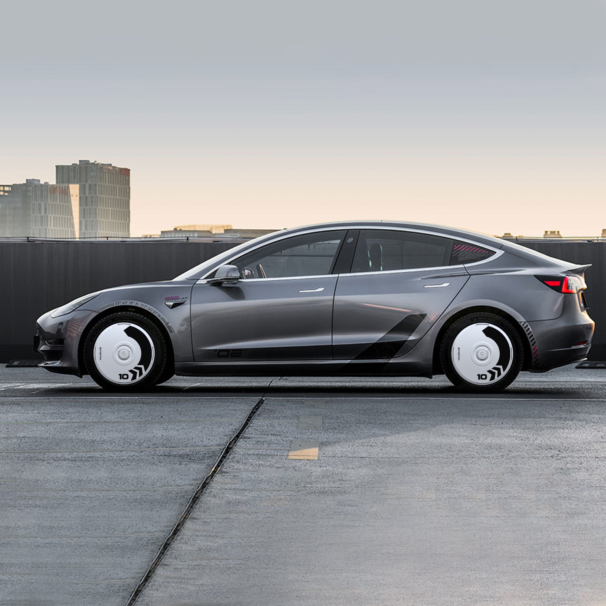 A Tesla Model 3 (2021-2023) with HALOBLK Store’s HALODISC 2 Arrow Wheel Covers for 20" Überturbine wheels is parked on a rooftop, showcasing the stylish gray car and its distinctive white, futuristic hubcaps against a city backdrop.