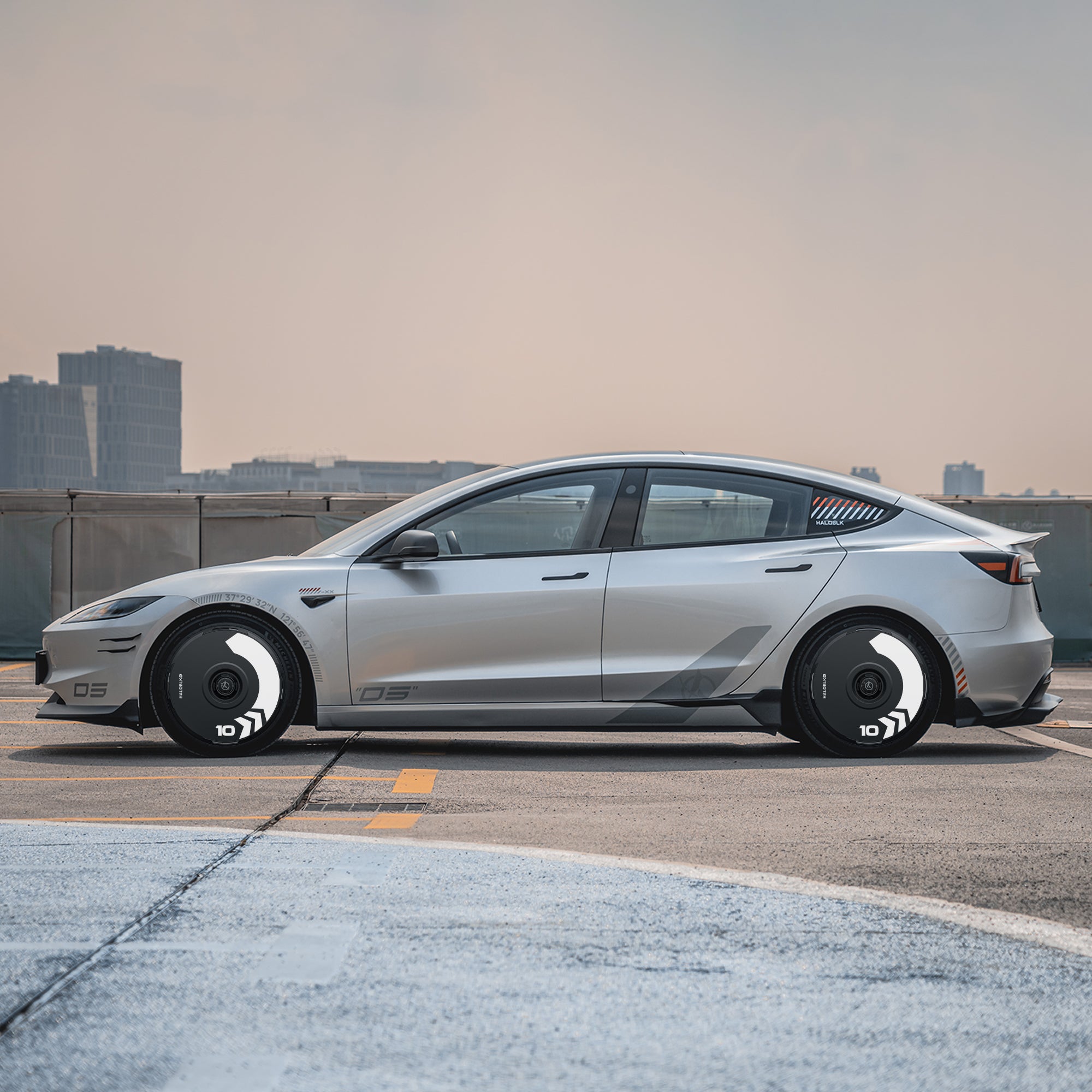 A Tesla Model 3 Highland with HALODISC 2 Arrow 19" wheel covers from HALOBLK Store is parked on a rooftop lot, showcasing its sleek silver design and stylish black-and-white wheels against a cityscape under a hazy sky.