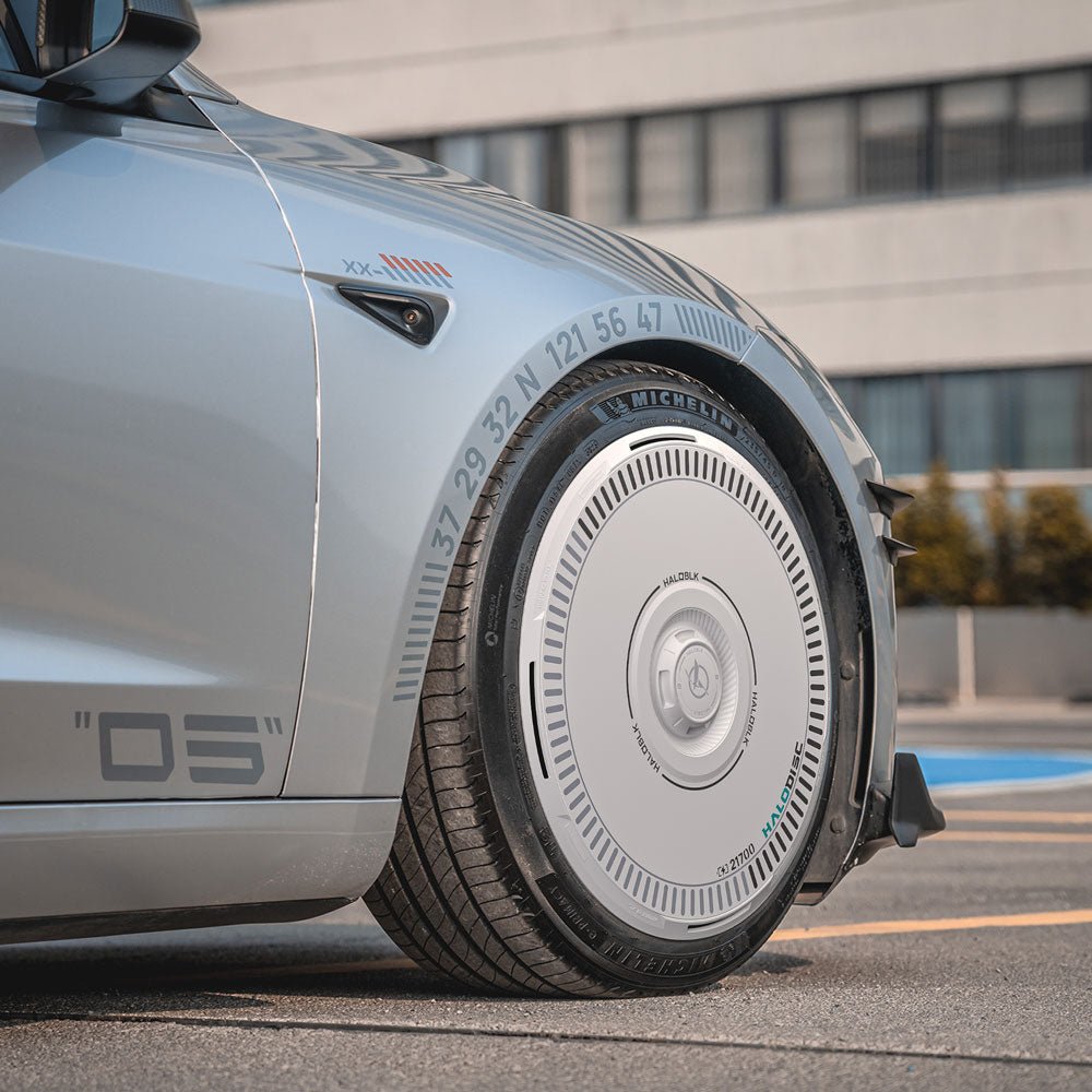 Close-up of HALODISC 2 Wheel Covers by HALOBLK Store on a Tesla Model 3 Highland 18" Photon wheel, showing the unique disc-shaped rim with numeric markings. The car is parked outdoors near a modern building with horizontal windows.