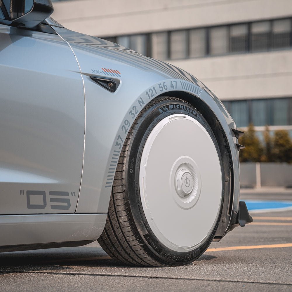 A close-up of a Tesla Model 3 Highland’s front wheel fitted with HALODISC 2 Wheel Covers (HALOBLK Store), showing a solid white aerodynamic cover with printed numbers and coordinates. A modern building and greenery appear in the background.