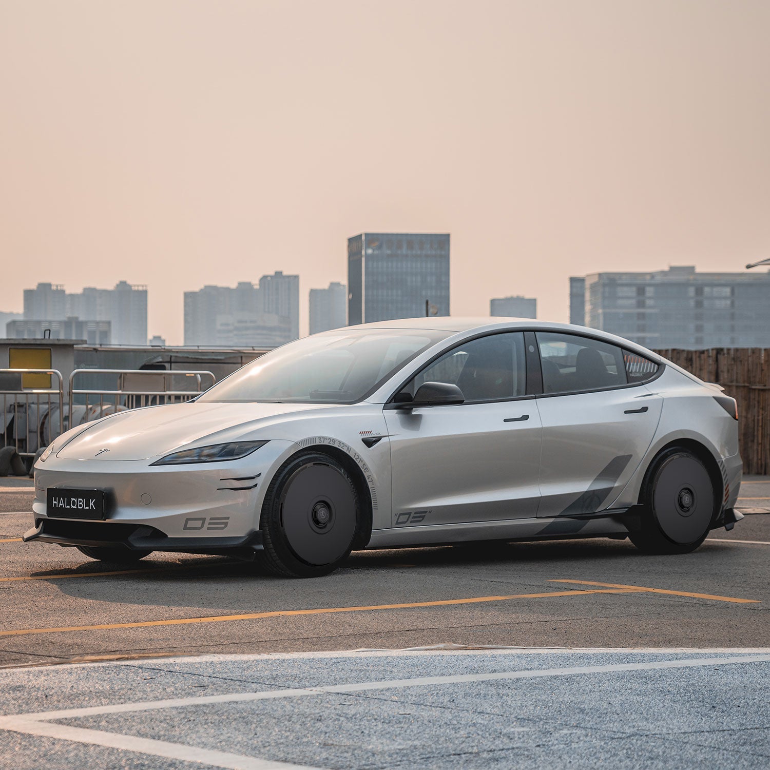 A Tesla Model 3 Highland with HALODISC 2 Wheel Covers from HALOBLK Store is parked on a rooftop, featuring black aerodynamic covers for the 20" Warp Wheels, silver finish, and dark tinted windows against a city skyline under a hazy sky.
