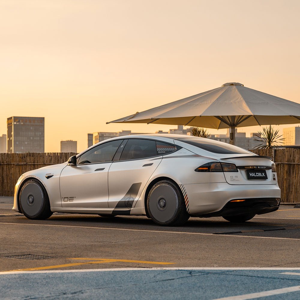 A silver Tesla Model S with HALODISC 2 Wheel Covers by HALOBLK Store is parked in an outdoor lot at sunset, modern city buildings behind it and a large white umbrella nearby. The car features sleek lines and covered wheels.