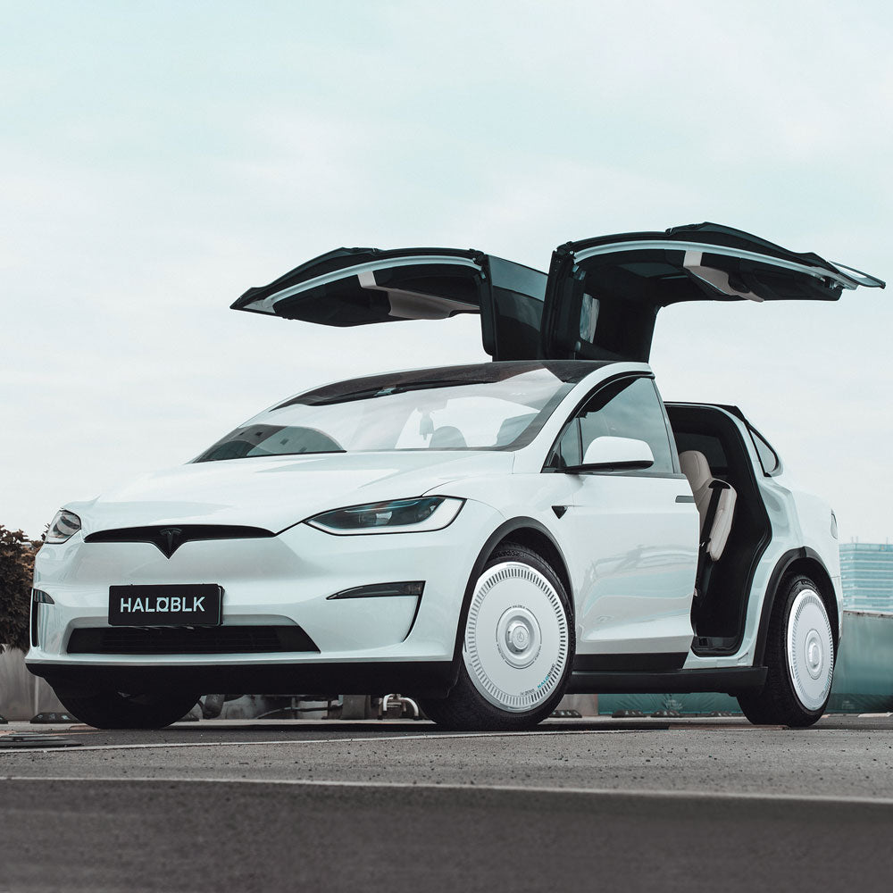 A white Tesla Model X (2016-2020) with HALODISC 2 Wheel Covers by HALOBLK Store on 20" Slipstream wheels, parked on concrete with falcon-wing doors open and front license plate reading HALOBLK under a cloudy sky.