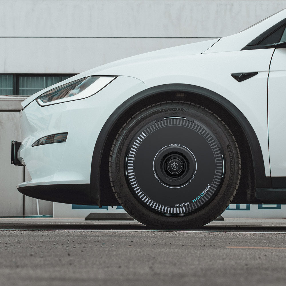 Close-up side view of a white Tesla Model X (2016-2020) with HALODISC 2 Wheel Covers by HALOBLK Store, featuring futuristic black covers with concentric white markings, parked on pavement in front of a white building.