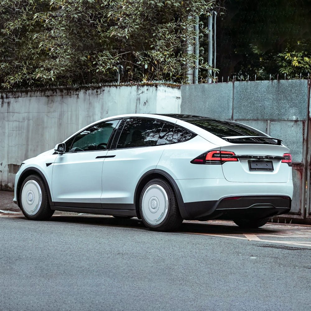 A white Tesla Model X with a black roof is parked by a high wall and greenery, viewed from the rear-side, and features HALODISC 2 Wheel Covers for 2021-2025 Model X 20" Cyberstream Wheels from HALOBLK Store.
