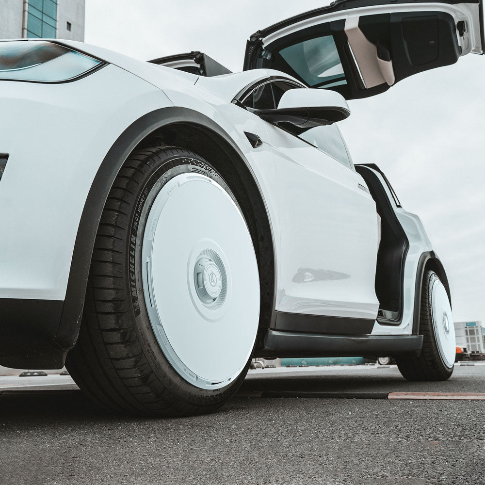 A low-angle view of a white Tesla Model X (2021-2025) with gullwing doors open, featuring HALODISC 2 Wheel Covers for 20" Cyberstream wheels by HALOBLK Store, parked near a modern building on asphalt.