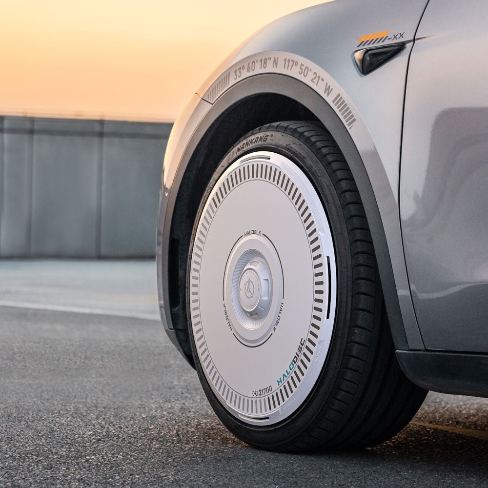 Close-up of the HALODISC 2 Wheel Covers for Tesla Model Y 20" Induction Wheels by HALOBLK Store, showing their smooth, white aerodynamic design with fine markings on a silver wheel, parked on pavement at sunset.