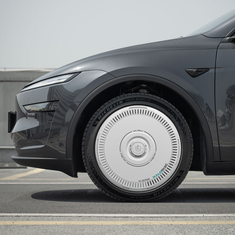 A close-up side view of a gray Tesla Model Y with HALODISC 2 Wheel Covers by HALOBLK Store on 20" Helix 2.0 wheels, featuring large white disc-shaped hubcaps, parked on a road with a blurred building in the background.